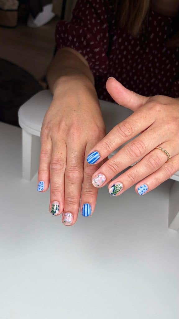 Woman's hands display blue-themed nail art with stripes and floral designs on a clear base, showcasing a coordinated manicure.